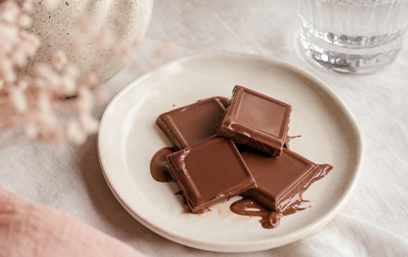 Squares of plain milk chocolate softening on a cream ceramic plate on a pale linen table beside a glass of water