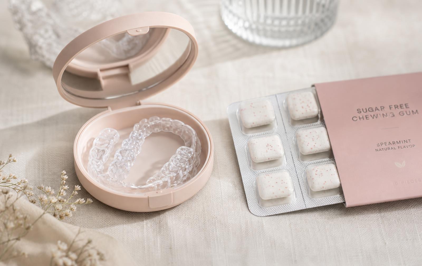 A clear plastic aligner tray in a blush case beside a blister pack of sugar free chewing gum on pale linen