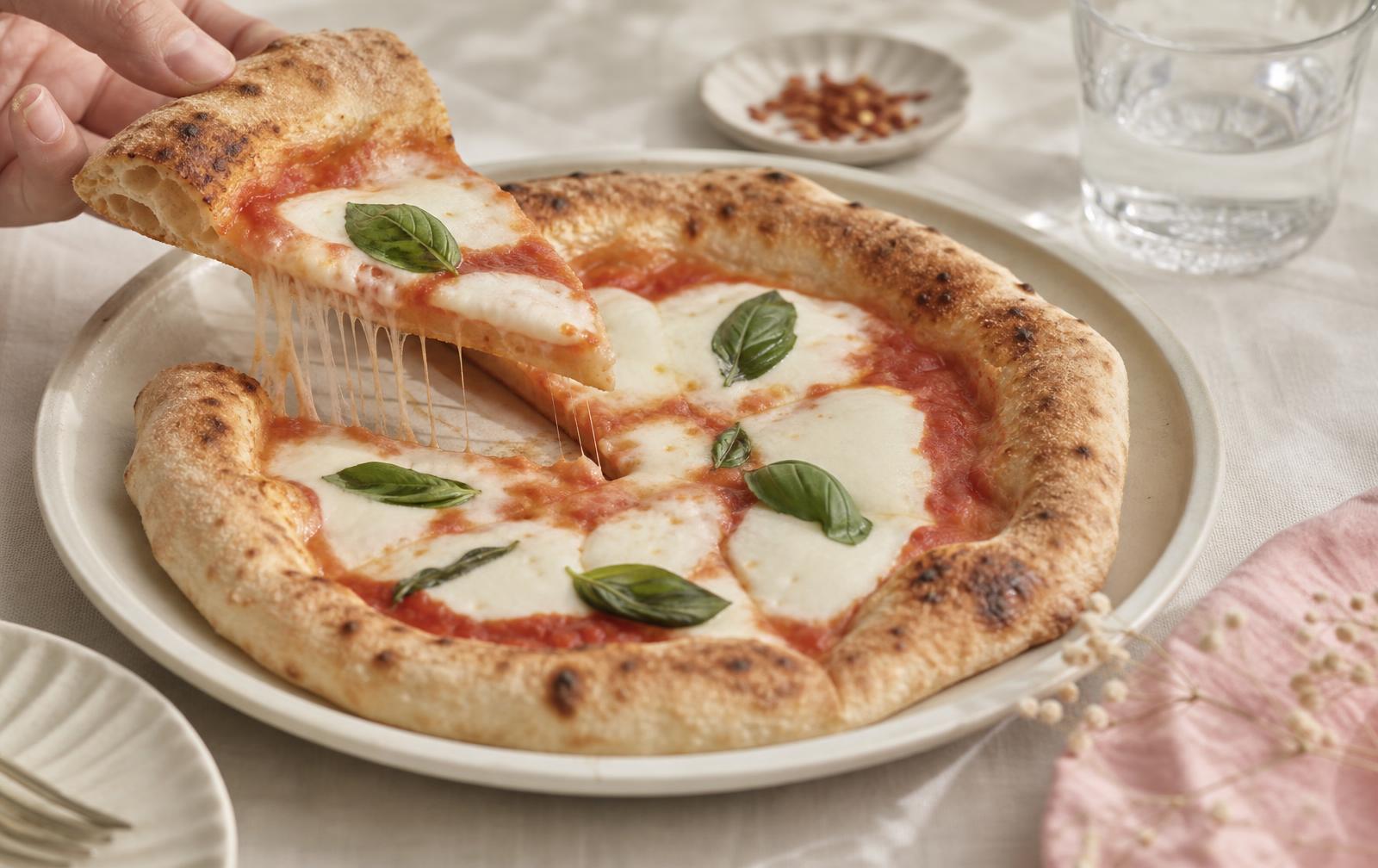 A margherita pizza on a light ceramic plate with one soft slice being lifted, on a pale linen table