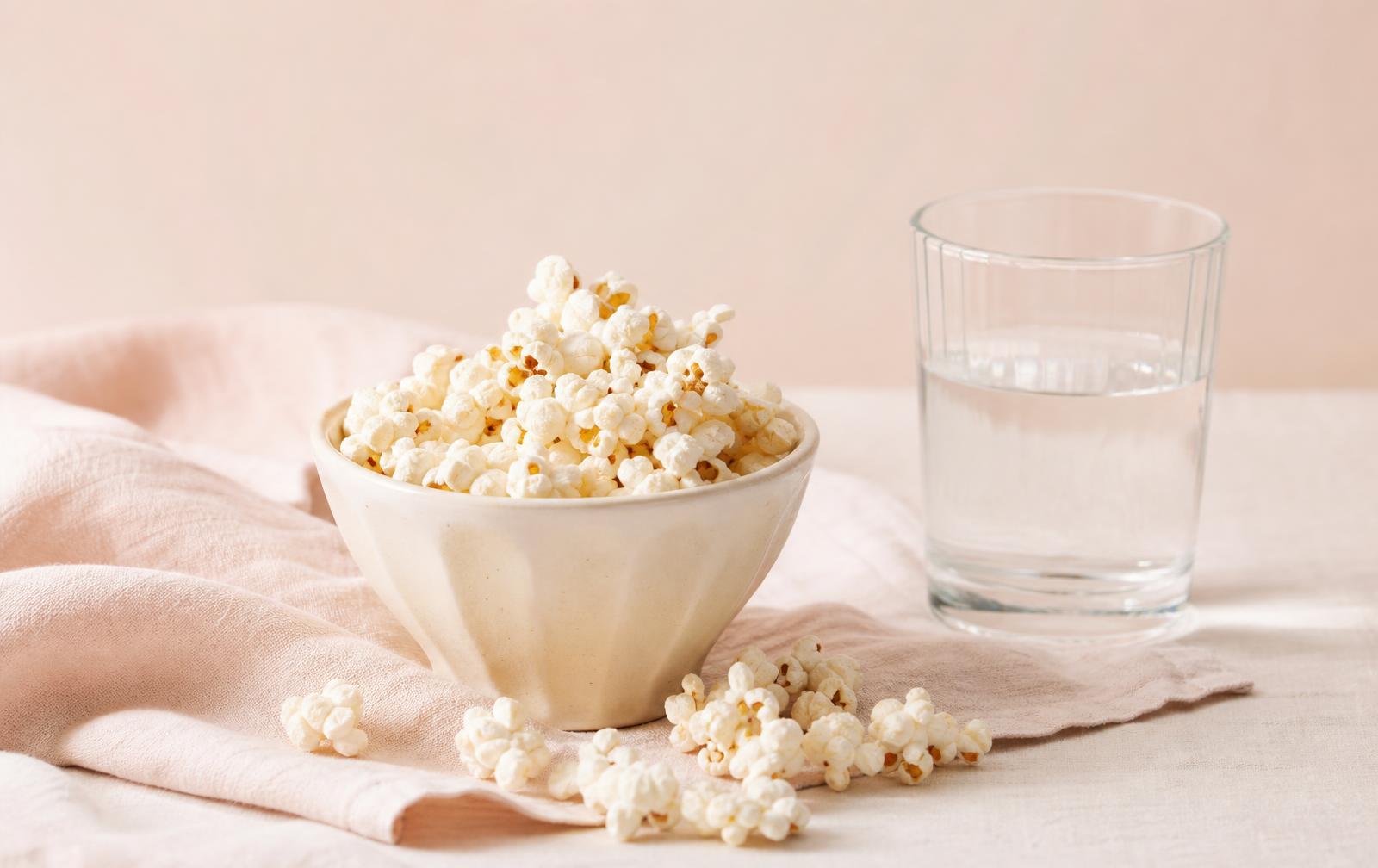 A cream ceramic bowl of freshly popped popcorn spilling onto a pale linen surface beside a glass of water