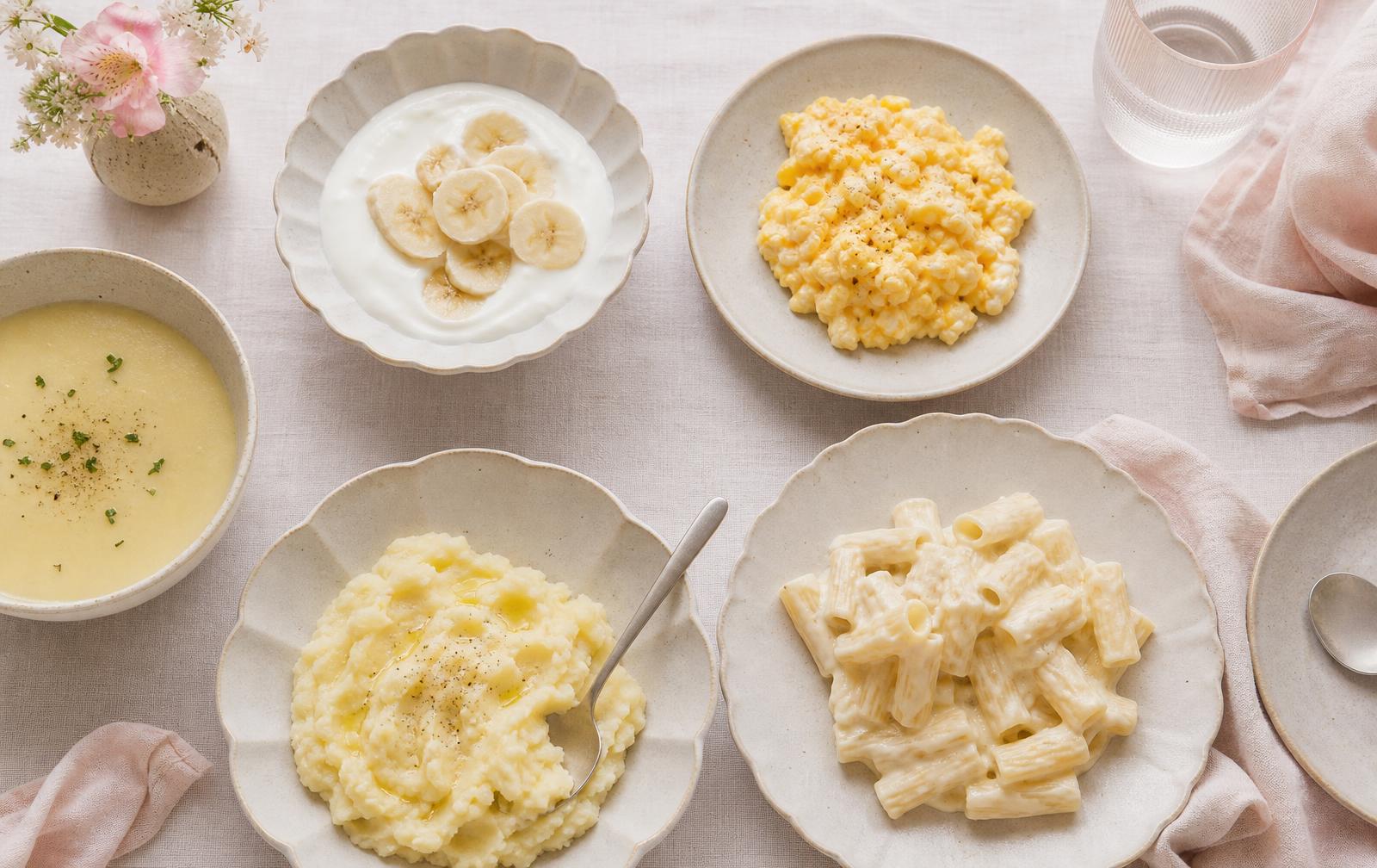 An overhead spread of soft braces friendly foods on cream plates including yoghurt with banana, scrambled eggs, mashed potato, soup and soft pasta