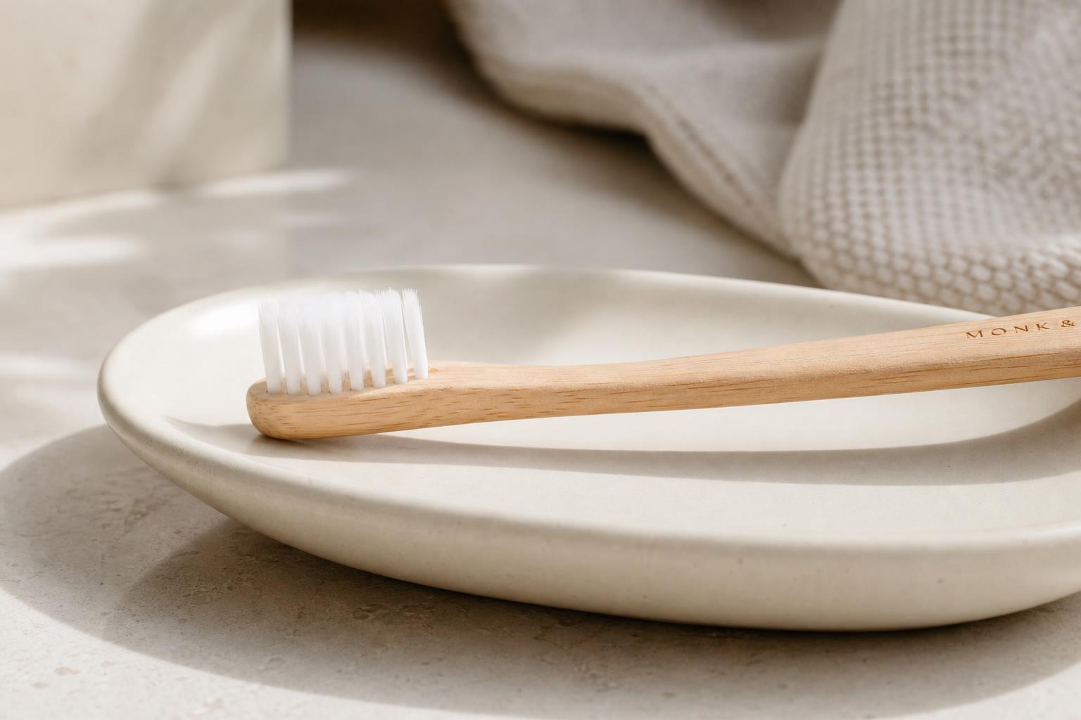 Soft bamboo toothbrush resting on a cream ceramic dish beside a linen towel in soft daylight